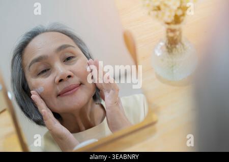 Attraktive ältere Erwachsene im mittleren Alter 50 Jahre alte asiatische Frau, die pflegende Antiage Gesichtshaut-Pflege-Creme-Behandlung anwendet, Blick auf den Spiegel, der täglich morgens tut Stockfoto