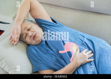 Ältere Frau, die auf dem Sofa schläft und unter starken Magenschmerzen leidet, ihren Bauch zu Hause unangenehm hält. Versorgungskonzept für ältere Menschen. Stockfoto