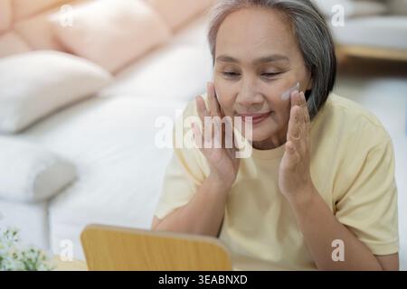 Reife asiatische Frau mit kurzen grauen Haaren sitzt am Tisch und legt ihr im Wohnzimmer Creme auf das Gesicht. Hautpflege und Schönheitsroutine für alternde Ski Stockfoto