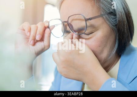 Eine gestresste Geschäftsfrau, die an Kopfschmerzen und Augenmüdigkeit leidet, reibt sich an den Schläfen und hält eine Brille, während sie an ihrem Schreibtisch sitzt. Stockfoto