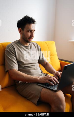 Ein Mann in lässiger Kleidung arbeitet von zu Hause aus mit einem Laptop. Er sitzt auf einer leuchtend gelben Couch, fokussiert auf den Bildschirm. Moderne Einrichtung mit Tageslicht. Konzept von Stockfoto