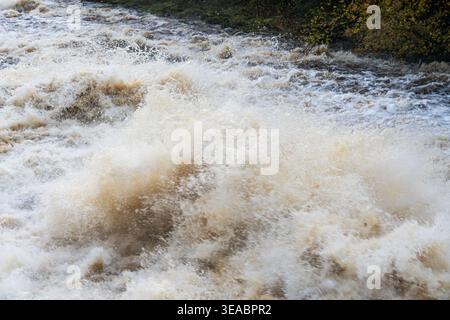 Turbulentes, torfbraunes Hochwasser, das in einem Nahaufnahme-Abstract eines aufgeschwollenen Flusses in Wasserflut zusammenbricht und schäumt Stockfoto
