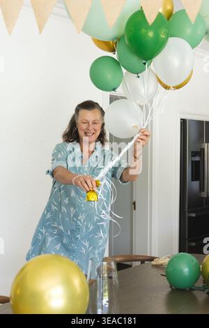 Ältere Frau, die am Tisch in der Küche steht, in blauem Kleid mit grünen Weißgoldballons Stockfoto
