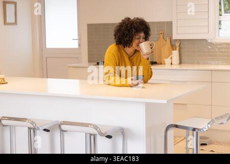 Afroamerikanerin im Senfstrick, lehnt sich auf die weiße Kücheninsel, schlürft einen Becher, überprüft das Telefon Stockfoto