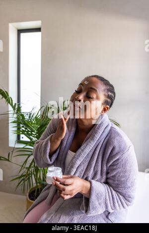Afroamerikanerin mittleren Alters, die im Badezimmer Gesichtscreme aus dem offenen Glas in einen Bademantel aufträgt Stockfoto