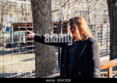 Junge kaukasische Frau, die mit überraschtem Ausdruck in der Nähe von Tiergehege im Zoo mit Zaun und Bäumen bei Tageslicht nach vorne zeigt. Emotion, Reaktion Stockfoto