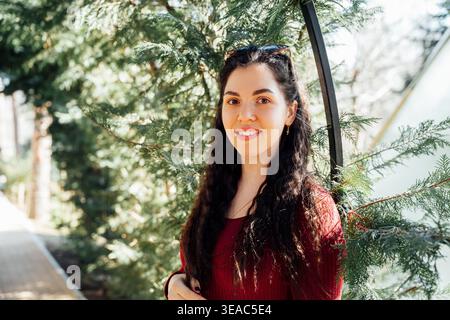 Junge kaukasische Frau mit langen dunklen Haaren, die einen roten Pullover trägt und in einem Park neben grünen Nadelbaumzweigen steht und in die Kamera blickt. Hochformat, außen Stockfoto