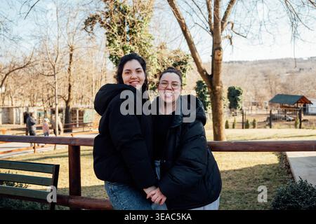 Zwei kaukasische Schwestern stehen zusammen und halten Hände in einem Zoo, während sie lächeln und bei Tageslicht posieren, mit Bäumen und Zaun im Hintergrund. Familie Stockfoto