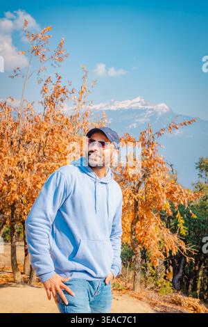Ein Mann in einem blauen Hoodie, der vor Bäumen mit gelben Blättern in der Nähe des Tungnath Tempels in Chopta, Uttarakhand, Indien steht. Die malerische Aussicht auf den Berg Stockfoto