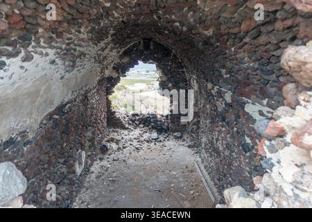 Ein Blick aus einer gewölbten Steinkammer auf die Ruinen der Burg Akrotiri mit Blick durch eine zerklüftete Öffnung in Richtung der Landschaft von Santorin. Stockfoto