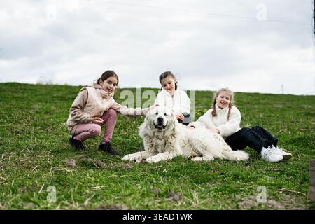 Drei glückliche Mädchen sitzen auf einem grasbewachsenen Hügel, streicheln einen großen weißen Hund und genießen das Spielen im Freien. Stockfoto
