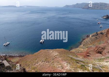 Blick auf die luxuriösen Katamarane vor Anker in der tiefblauen Ägäis, eingerahmt von den roten vulkanischen Hängen der Santorini-Caldera in der Nähe von Oia. Stockfoto