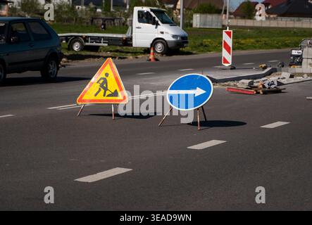 Verkehrsschilder mit Straßenreparaturen und Umweg, die Fahrzeuge an einer Baustelle auf einer Asphaltstraße mit geparktem Auto vorbeiführen Stockfoto