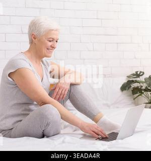Ältere Frau, die tagsüber auf dem Bett sitzt und ihr Laptop benutzt Stockfoto