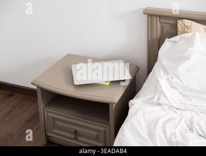 Geschlossener Laptop auf einem Nachttisch neben einem Bett mit zerknitterter weißer Bettwäsche, die Technologie in entspannter Atmosphäre darstellt Stockfoto