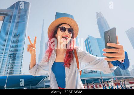 Frau, die ein Reiseselfie in Dubai macht, in die Smartphone-Kamera lächelt und ein Friedenszeichen zeigt Stockfoto