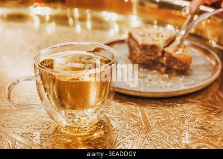 Tee in einer Glasschale und eine Person, die ein Stück Baklava von einem Teller auf einem kunstvollen goldenen Tablett nimmt. Traditionelles Dessert Stockfoto