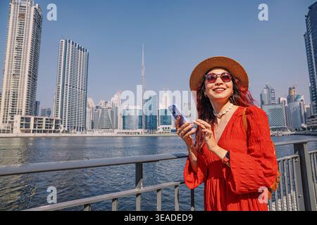 12. Januar 2026, Dubai, VAE: Frau in rotem Kleid und Hut, die die Uferpromenade von Dubai genießt und ein Smartphone mit dem Burj Khalifa im Hintergrund hält Stockfoto