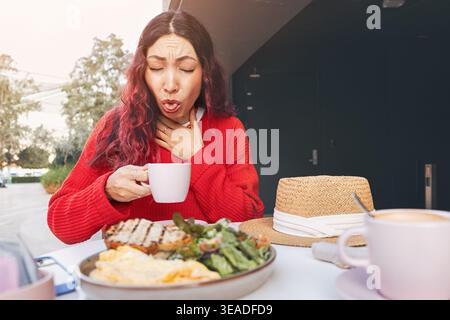 Junge Frau, die Schmerzen und Ersticken beim Trinken von heißem Kaffee in einem Restaurant im Freien während der Frühstückszeit hat Stockfoto