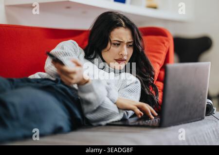 Junge Frau auf der Couch mit Laptop und Fernbedienung, die frustriert aussah, während sie vom Sofa aus surfte Stockfoto
