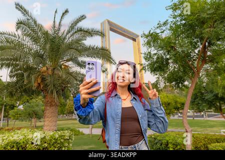 Junge Frau mit rotem Haar posiert für ein Selfie-Video mit Smartphone, mit Gesten Friedensschild in der Nähe von Dubai Frame. Reise- und Tourismuskonzept Stockfoto