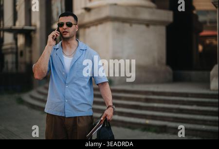 Mann, der am Telefon spricht, in lässigem blauem Hemd und Sonnenbrille, der mit einer Tasche an der Stadttreppe vorbeigeht Stockfoto