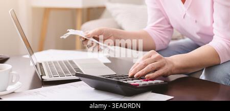 Die Person arbeitet zu Hause mit Rechner und Laptop, wobei die Papiere auf dem Tisch liegen, während sie Quittungen hält Stockfoto