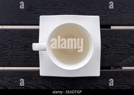 Blick von oben auf eine leere weiße Tasse auf einer quadratischen weißen Untertasse, die auf einem dunklen Lattentisch steht. Selektiver Fokus, Kopierraum Stockfoto