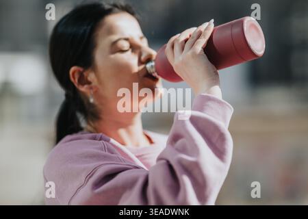 Junge Frau trinkt draußen aus einer wiederverwendbaren Wasserflasche in lässigem rosa Sweatshirt Stockfoto