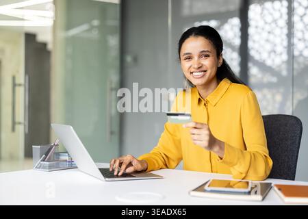 afroamerikanerin lächelt in die Kamera, hält eine Kreditkarte und tippt auf einem Laptop, macht Online-Shopping und sichere Zahlungstransaktionen aus einer modernen Büroumgebung Stockfoto