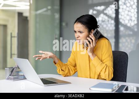 Frustrierte junge Geschäftsfrau, die ein wütendes Gespräch auf einem Mobiltelefon, einen Laptop benutzt, Stress und Probleme während der Arbeit in einem modernen Büro erlebt Stockfoto