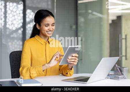 Eine junge Geschäftsfrau lächelt an einem digitalen Tablet an, während sie an einem Bürotisch mit Laptop arbeitet, und zeigt moderne Konnektivität, Produktivität und selbstbewussten beruflichen Erfolg Stockfoto