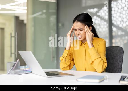 Eine gestresste junge Frau, die an ihrem Schreibtisch arbeitet und schwere Kopfschmerzen entwickelt, die Schläfen mit den Händen drückt, während sie Schmerzen, Burnout und emotionale Ermüdung durch ständigen Arbeitsdruck erlebt Stockfoto