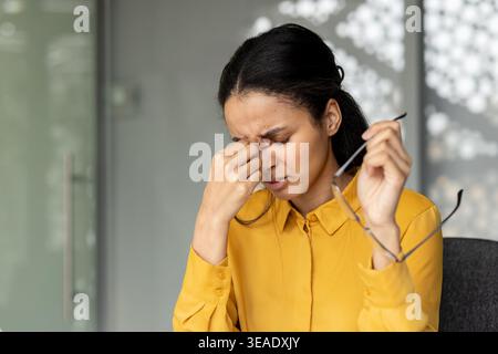 Junge Frau fühlt sich müde und gestresst, massiert ihren Nasensteg und entfernt Brillen, während sie im Büro unter Kopfschmerzen und Augenanstrengung leidet Stockfoto