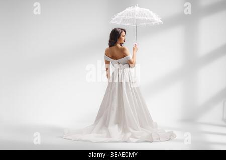 Braut in schulterfreiem Hochzeitskleid mit Spitzenschirm in einem Studio Stockfoto