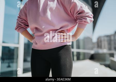 Selbstbewusste Frau in rosafarbenem Sweatshirt und schwarzer Leggings, die draußen mit Händen auf den Hüften steht Stockfoto