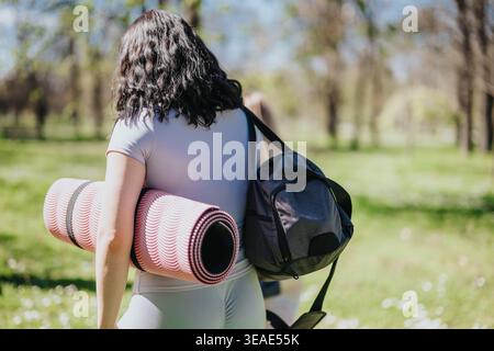 Frau mit Yogamatte und Sporttasche, die durch den sonnigen Park geht, bereit für Outdoor-Workout Stockfoto