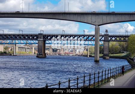 Die Aussicht zeigt die Redheugh Bridge, die den Fluss Tyne überspannt und Newcastle mit Gateshead verbindet. Die Szene fängt den belebten Fluss unten ein, wobei Wasser fließt und Bewegungen auf der Brücke darüber sichtbar sind. Fußgänger können auf dem Weg am Fluss gesehen werden. Der Tag ist klar und es gibt Wolken am Himmel. Stockfoto