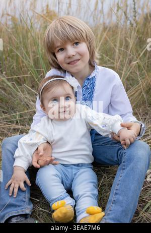 Baby Mädchen und Junge, 8-9 Jahre alt, Bruder Schwester, sitzt auf Gras und umarmt. Die Kinder tragen aufgestickte ukrainische Kleidung. Eng gestrickte Geschwister, f Stockfoto