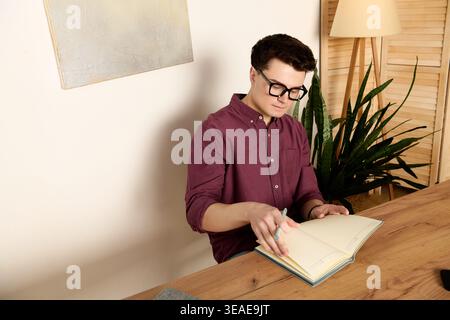Ein hübscher junger Mann in Brille schreibt in ein Notizbuch an seinem Holztisch zwischen Pflanzen. Stockfoto
