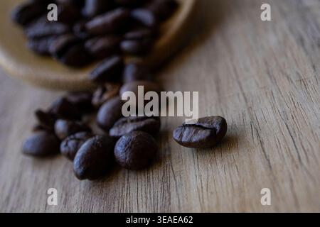 Nahaufnahme eines Lebensmittelfotos von gerösteten Kaffeebohnen, die aus einer Holzlöffel auf einer rustikalen Holzoberfläche mit geringer Schärfentiefe und weichem Licht verschüttet werden. Stockfoto