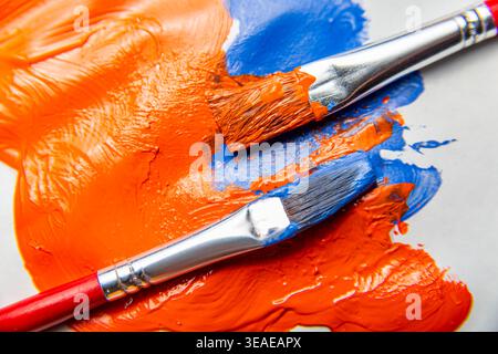 Zwei Pinsel sind mit orangefarbener und blauer Farbe bedeckt, während sie während einer kreativen Aktivität in einem Kunststudio auf einer weißen Oberfläche ruhen. Stockfoto