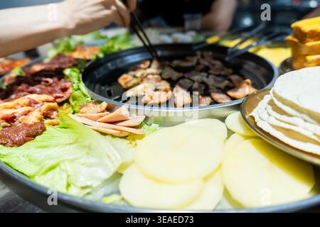 Nahaufnahme eines koreanischen Grillgerichts mit gegrilltem Fleisch, Kartoffelscheiben, Salatpackungen und Beilagen auf einem Stockfoto