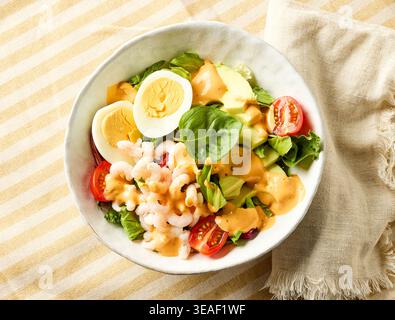 Frische gesunde Salatschüssel mit Garnelen, Avocadoscheiben, gekochtem Ei, Kirschtomaten und grünen Blättern, garniert mit cremigem Dressing, auf gestreifter Tischdecke Stockfoto