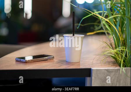Nahaufnahme einer leeren Kaffeetasse zum Mitnehmen und eines Mobiltelefons auf einem Holztisch. Moderner digitaler Lebensstil und nachhaltiges Verpackungskonzept mit grünen Pflanzen Stockfoto