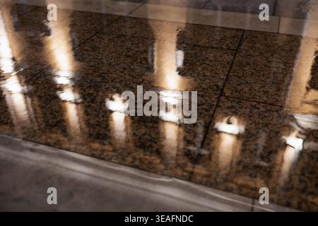 Abstrakte Lichter der Stadt spiegeln sich in einer Pfütze auf einem Bürgersteig aus Stein. Goldene Lichtreflexe auf nassen Granitfliesen bei Nacht. Stockfoto