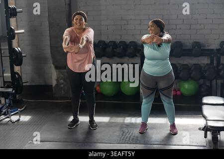 Vielfältige Freundinnen dehnen den Oberkörper in Sportbekleidung im Fitnessstudio auf Gummimatten von Hantelständer Stockfoto
