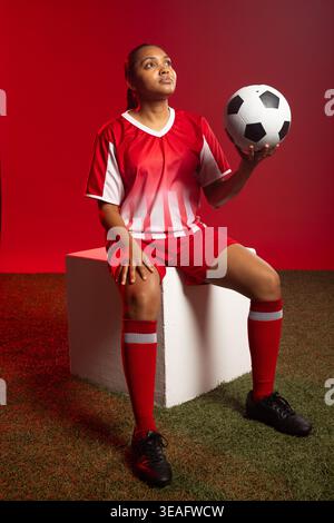 Fußballspielerin in rot-weiß-Uniform, die auf weißem Block auf dem Rasen sitzt und den Ball hält, der nach oben blickt Stockfoto