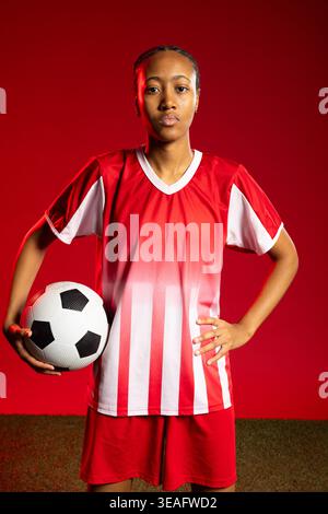 Afrikanische Fußballspielerin steht auf grünem Rasen rot in rot-weißem Trikot und hält Fußball Stockfoto