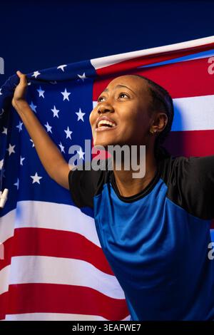 Afroamerikanerin, die die amerikanische Flagge hält und sie im Studio weit zieht, mit einem sportlichen Hemd Stockfoto